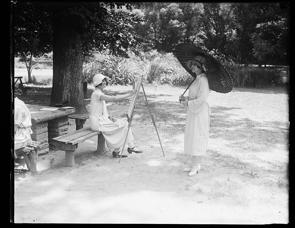 Black and white photograph of a young woman in 1920s dress painting another woman with a parasol.