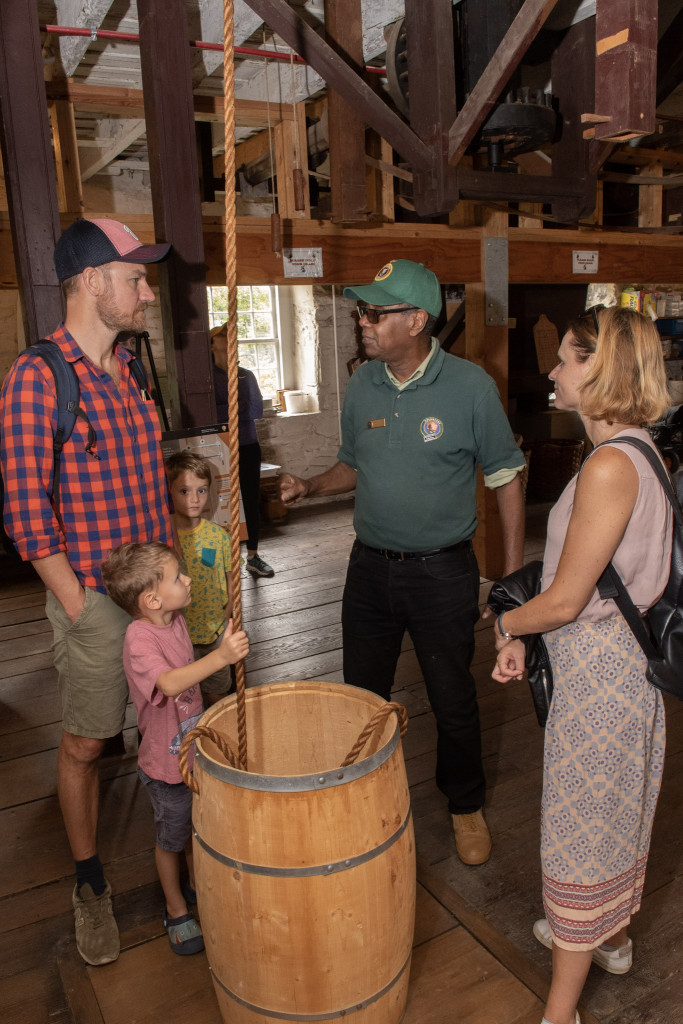 Man in a green NPS volunteer t-shirt shows a wooden barrel to Peirce Mill visitors.