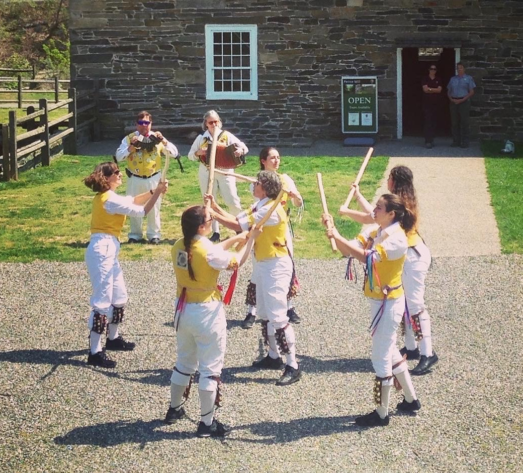 Women in yellow vests dance with sticks in front of an old stone building.