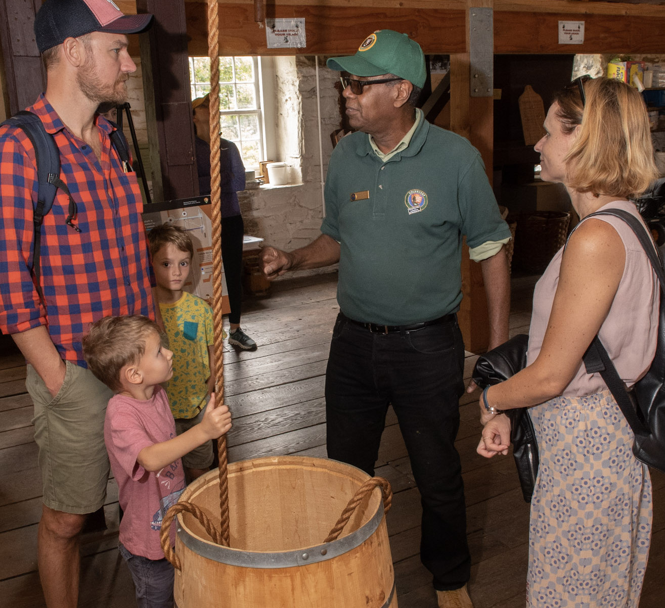 A man in a green NPS volunteer shirt explains the barrel hoist to mill visitors
