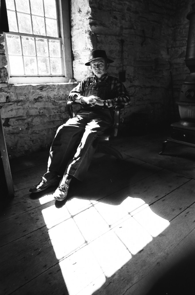 Man in an old-fashioned hat and overalls sitting next to a potbelly stove.