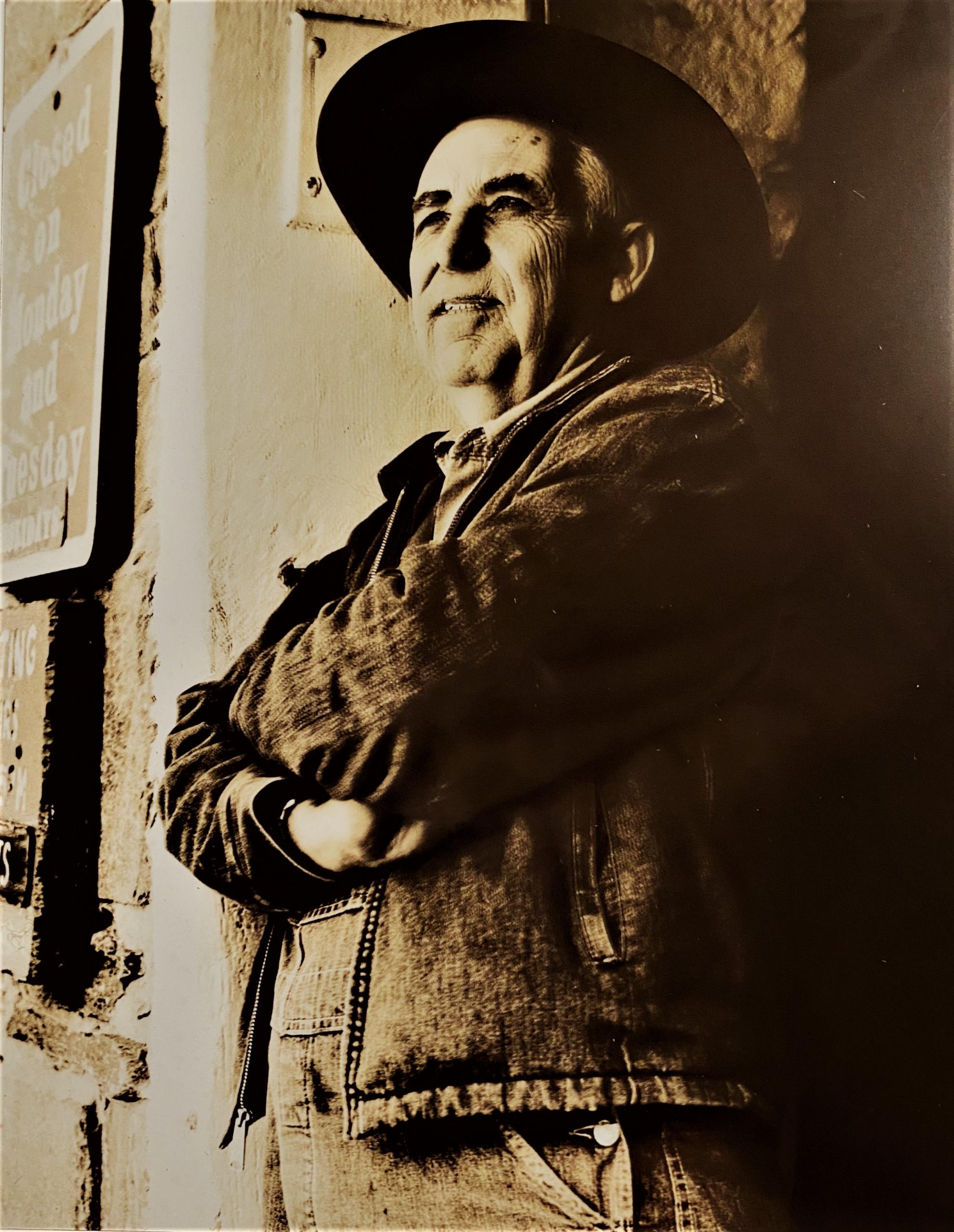 Black and white photograph of a man in brimmed hat and denim jacket leaning against the doorway of an old stone building