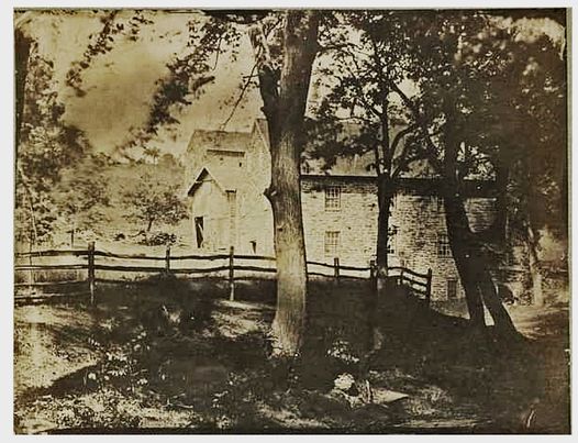 Old black and white photograph of stone buildings with trees in the foreground.