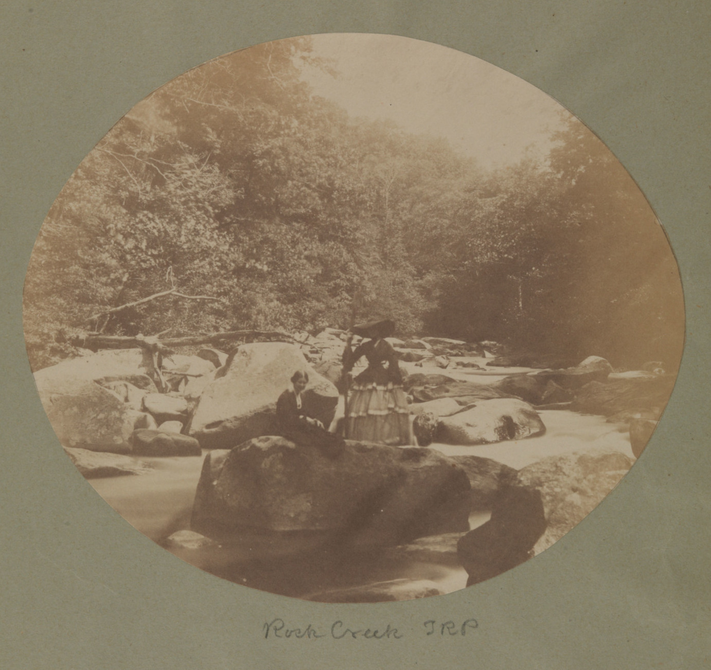 Very old black and white photograph of two women posing on large rocks in the middle of a creek.