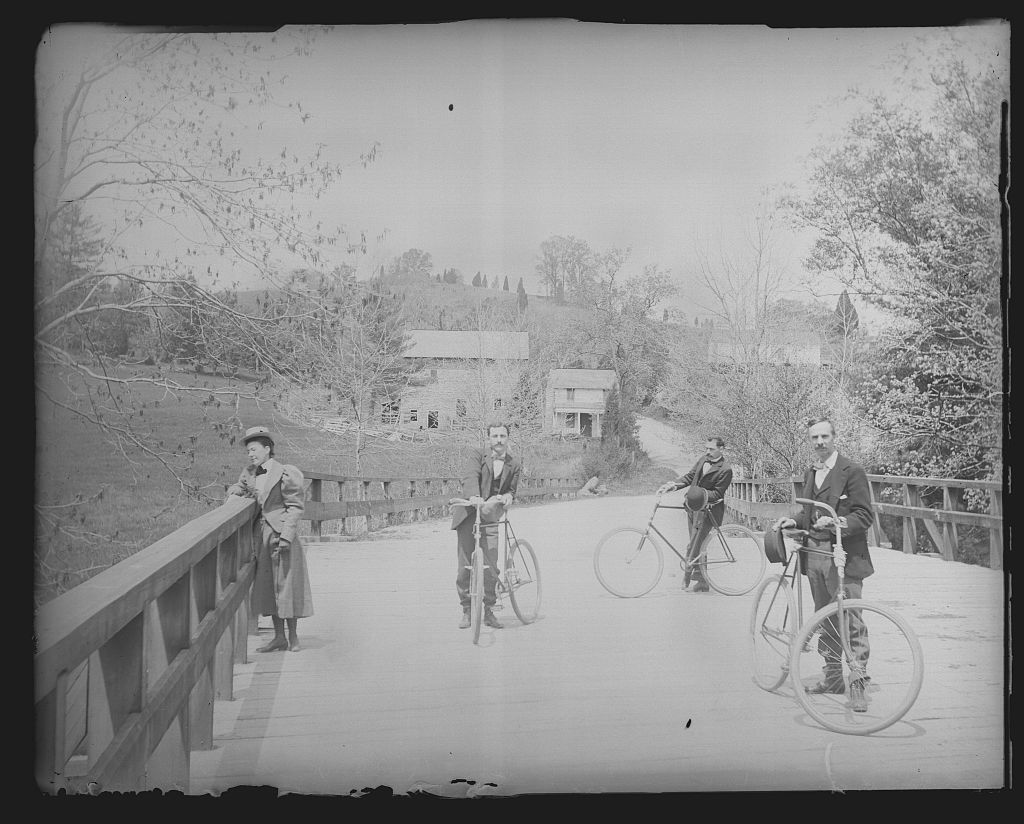Black and white photo of people standing on a bridge, some next to old- fashioned bicycles.
