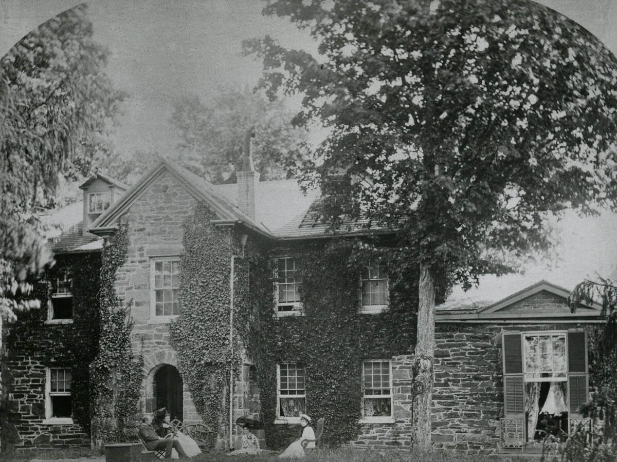 Old photograph of people sitting in front of a stone building covered in ivy.