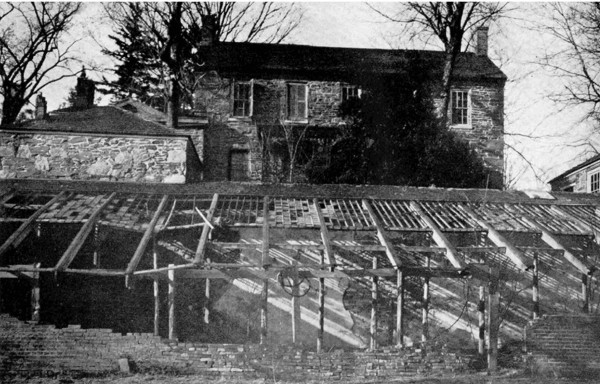 Black and white photograph showing the ruins of a greenhouse behind a large stone building.