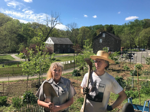 A woman in a park service uniform and a man in a straw hat stand in the middle of an orchard.