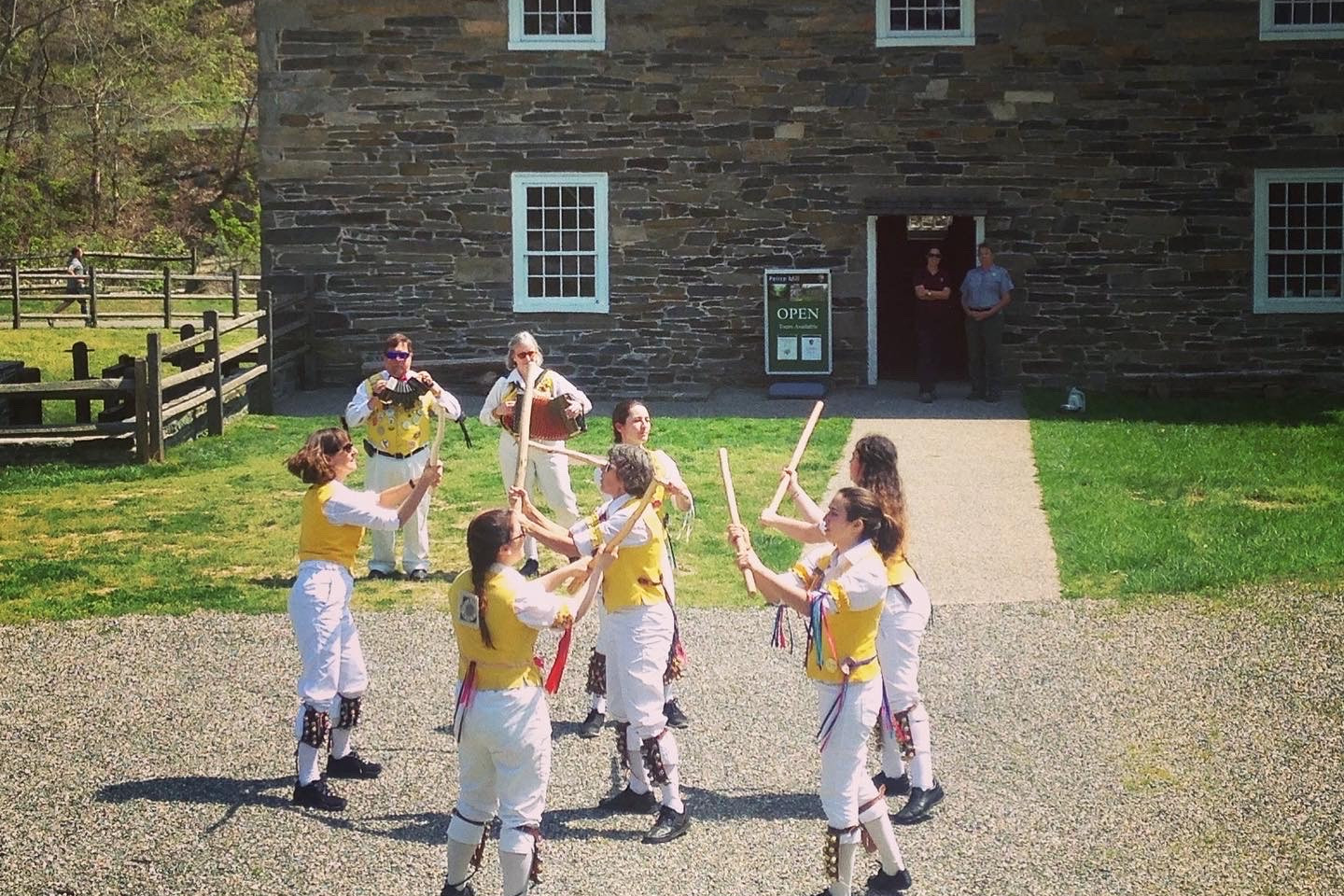 Morris Women 2022 Dancing Women in bright yellow vests and white knee pants dancing with sticks in front of Peirce Mill