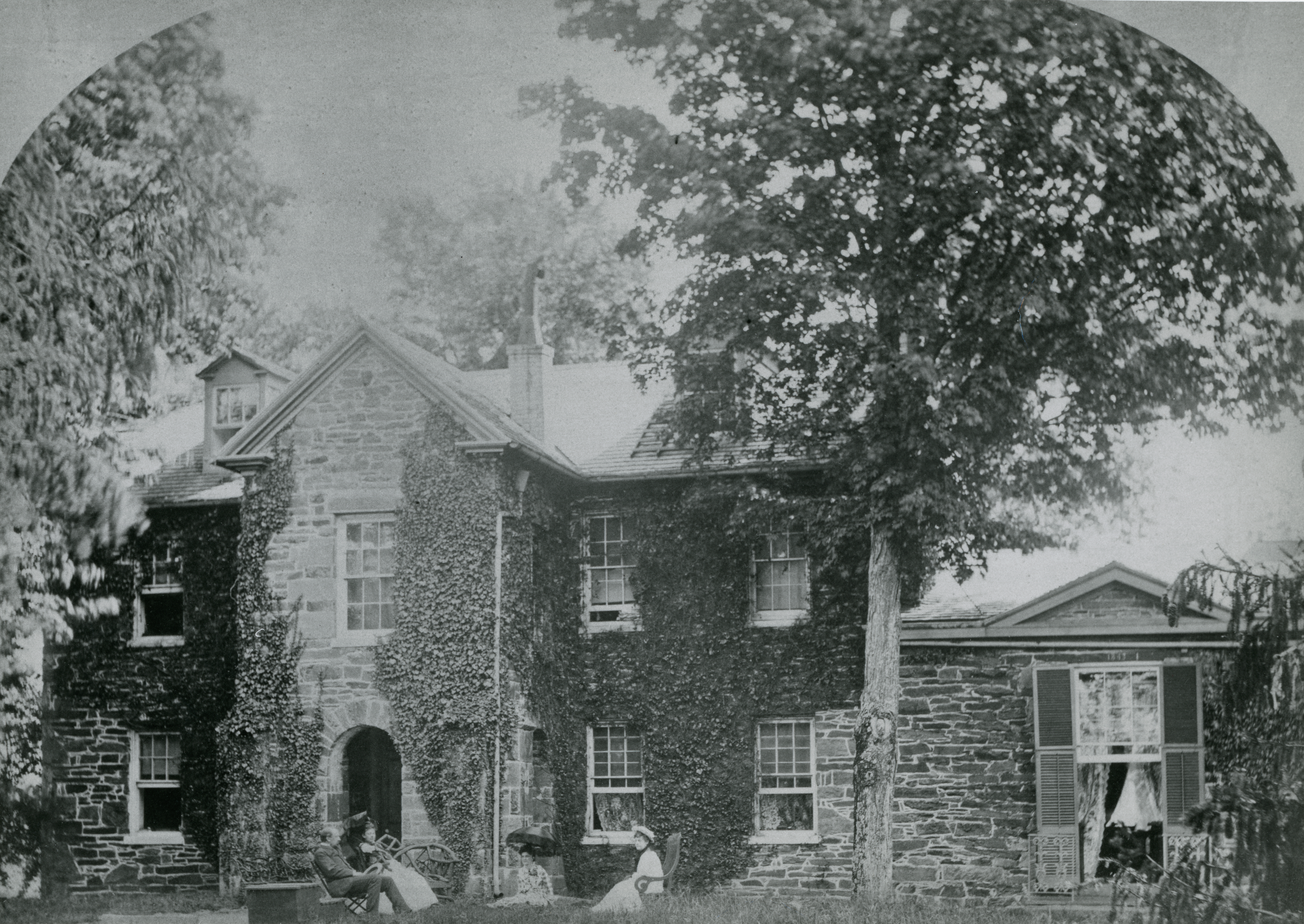 Photograph of a large stone mansion with two women and a man sitting in front of it.
