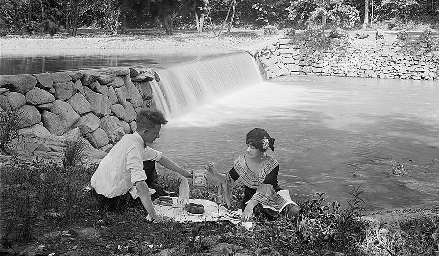 An early-twentieth century photograph of a man and woman picnicking in front of a waterfall.