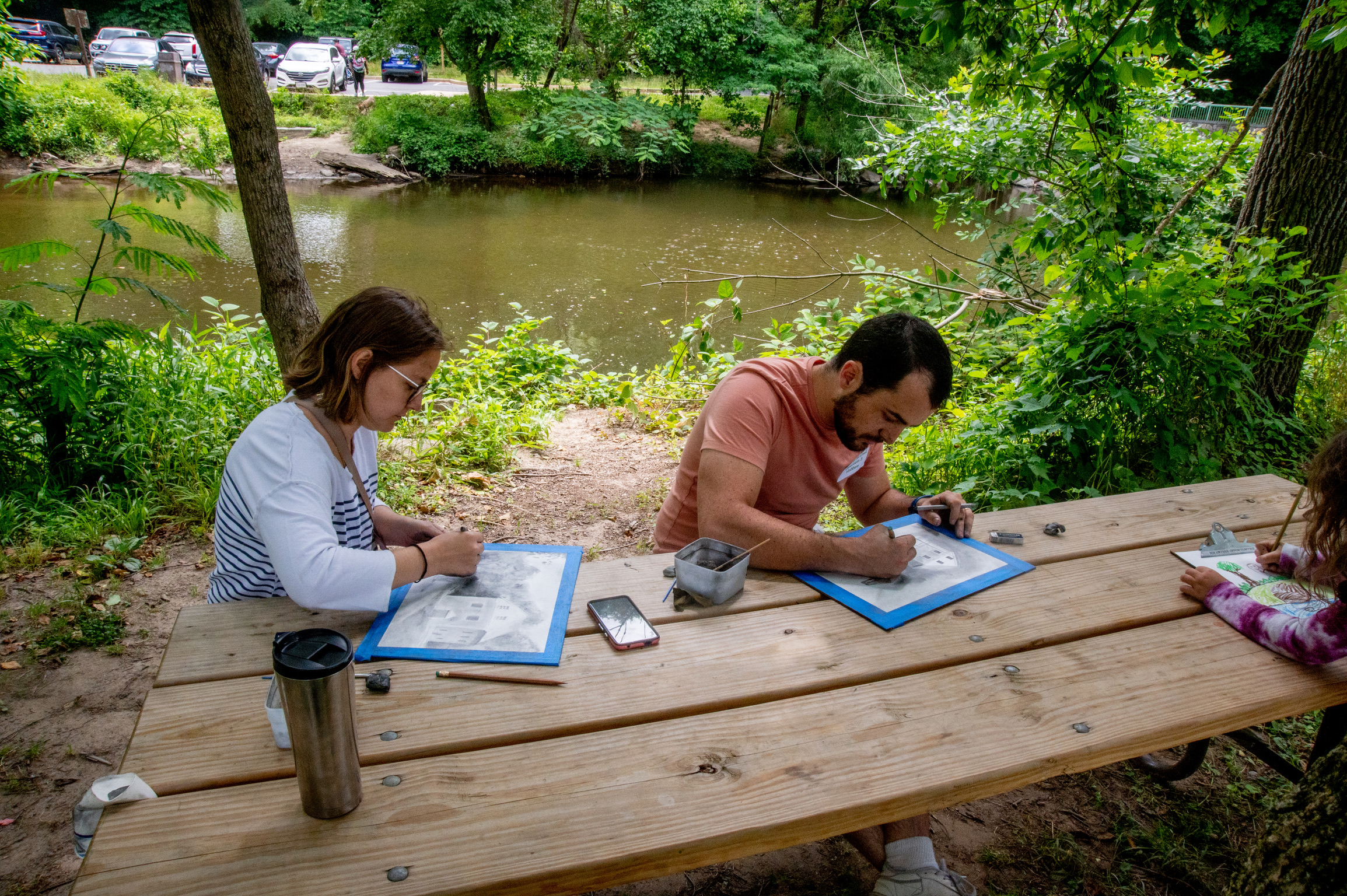 Two people draw at a picnic table beside Rock Creek.