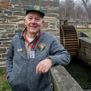 Photograph of a man in a National Park Service volunteer uniform standing next to a fence beside an old mill.