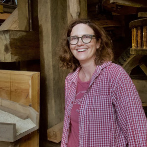 Photograph of a woman standing next to large wooden gears in the basement of a mill
